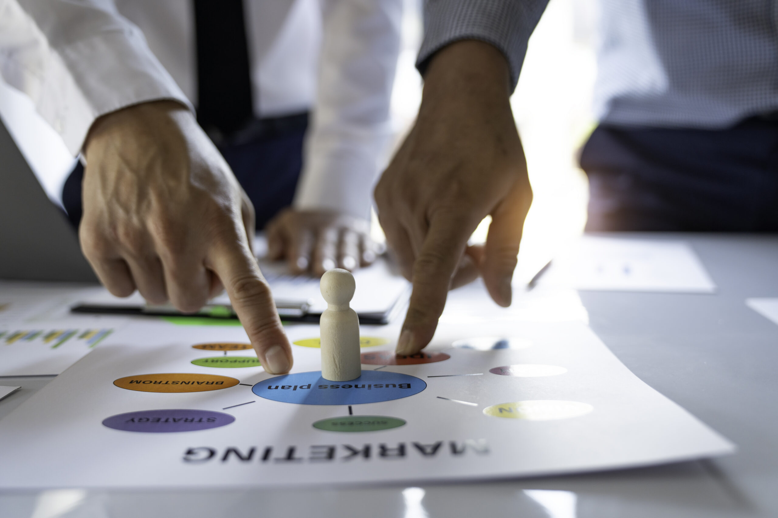 A wooden figure standing on a marketing solutions plan on a table, with two fingers pointing at the document.